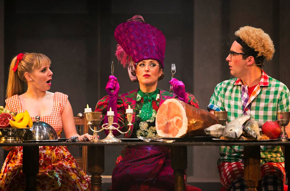 Actors Michelle Gallacher, Robert Jack and Dawn Sievewright are sat at a table wearing cheerful costumes designed by Alison Brown Costume Designer. Michelle wears purple and a large purple hat. She is holding up cutlery in front of a large ham.