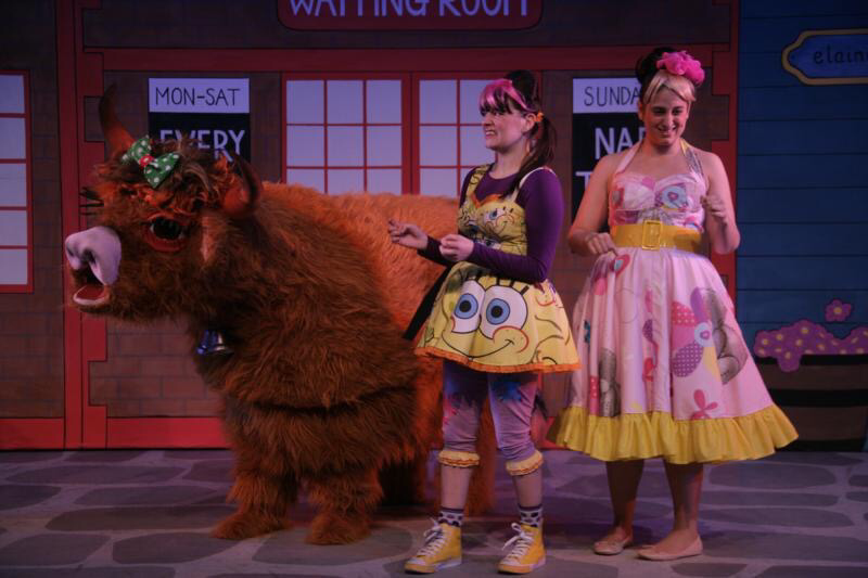 2 actors in panto wearing fun, colourful dresses are standing beside a panto Highland Cow. costumes designed by Alison Brown Costume Designer.